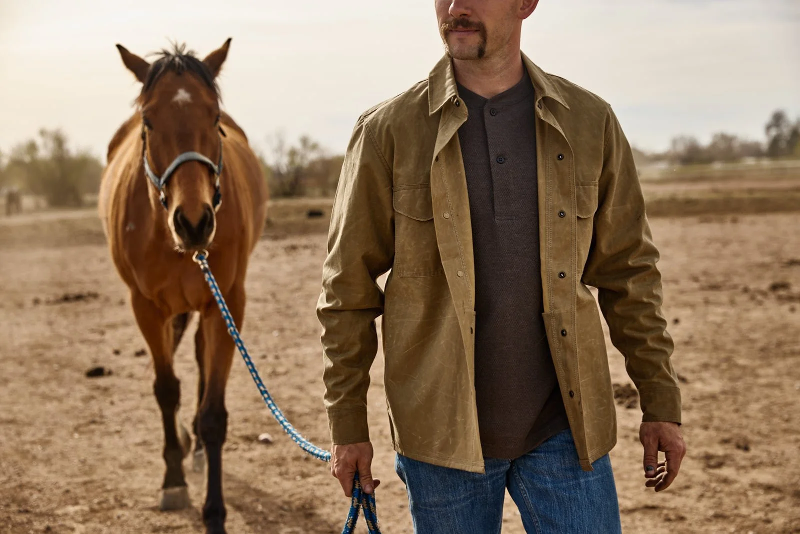 A man standing outdoors holding a blue rope lead, with a brown horse behind him on a dirt field.