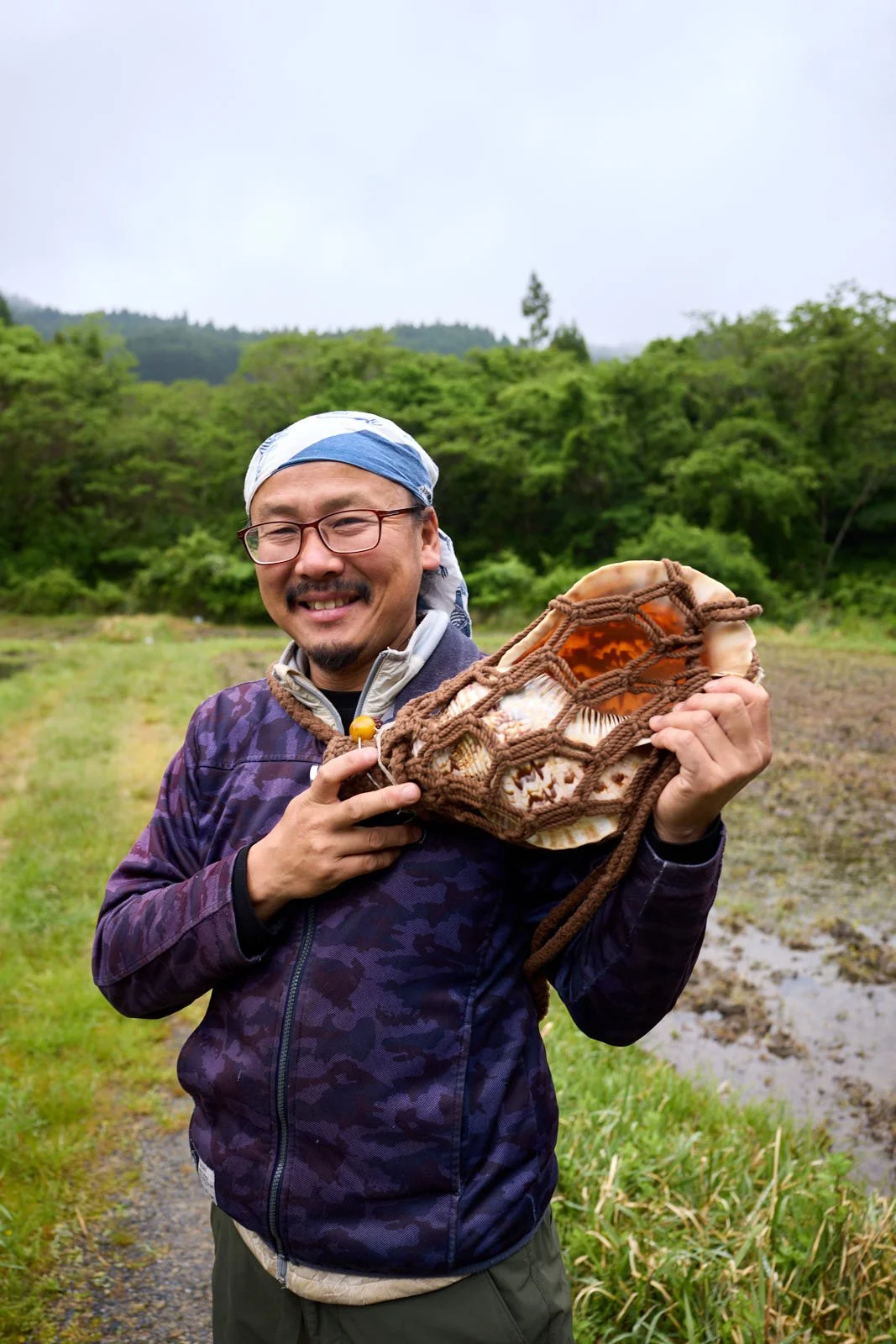 A smiling man wearing glasses, a bandana, and outdoor clothing, holding a conch shell, standing in a lush green field with trees in the background.