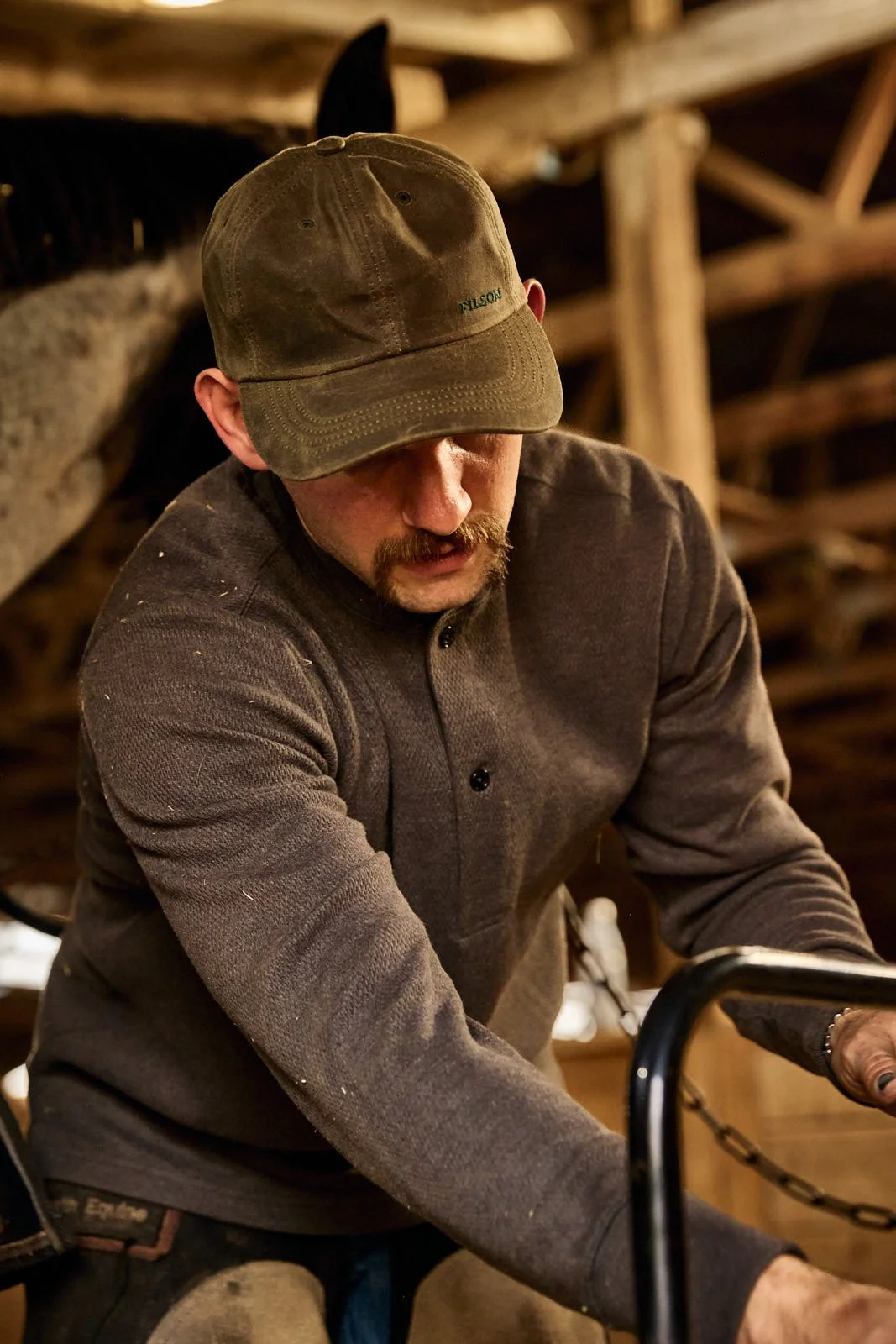 A man with a mustache wearing a green baseball cap and a long-sleeve gray shirt, working in a horse barn