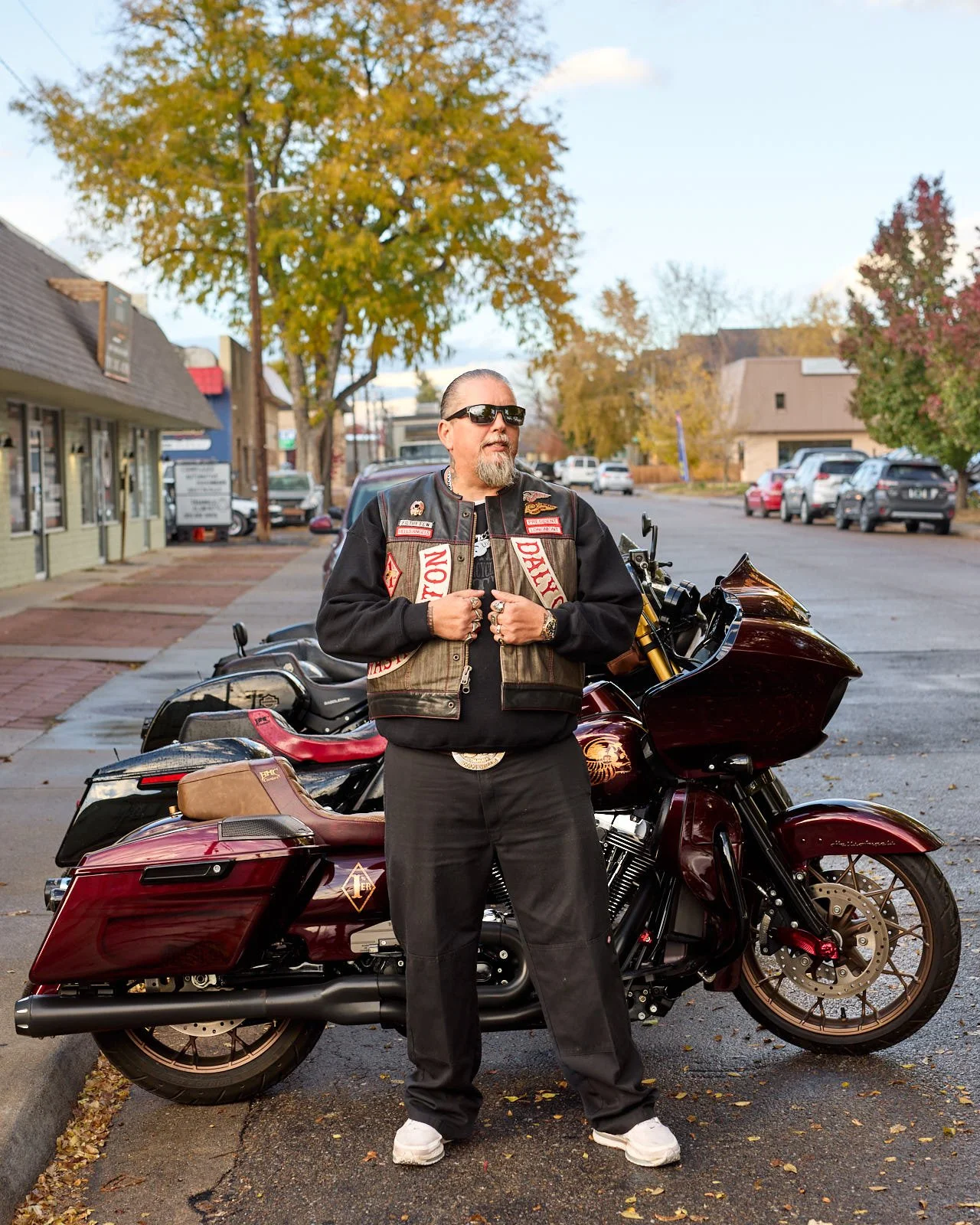 A man with sunglasses and a beard standing next to a maroon Harley Davidson motorcycle in a small town. The man wears a black jacket with patches and black pants, with trees and parked cars in the background.