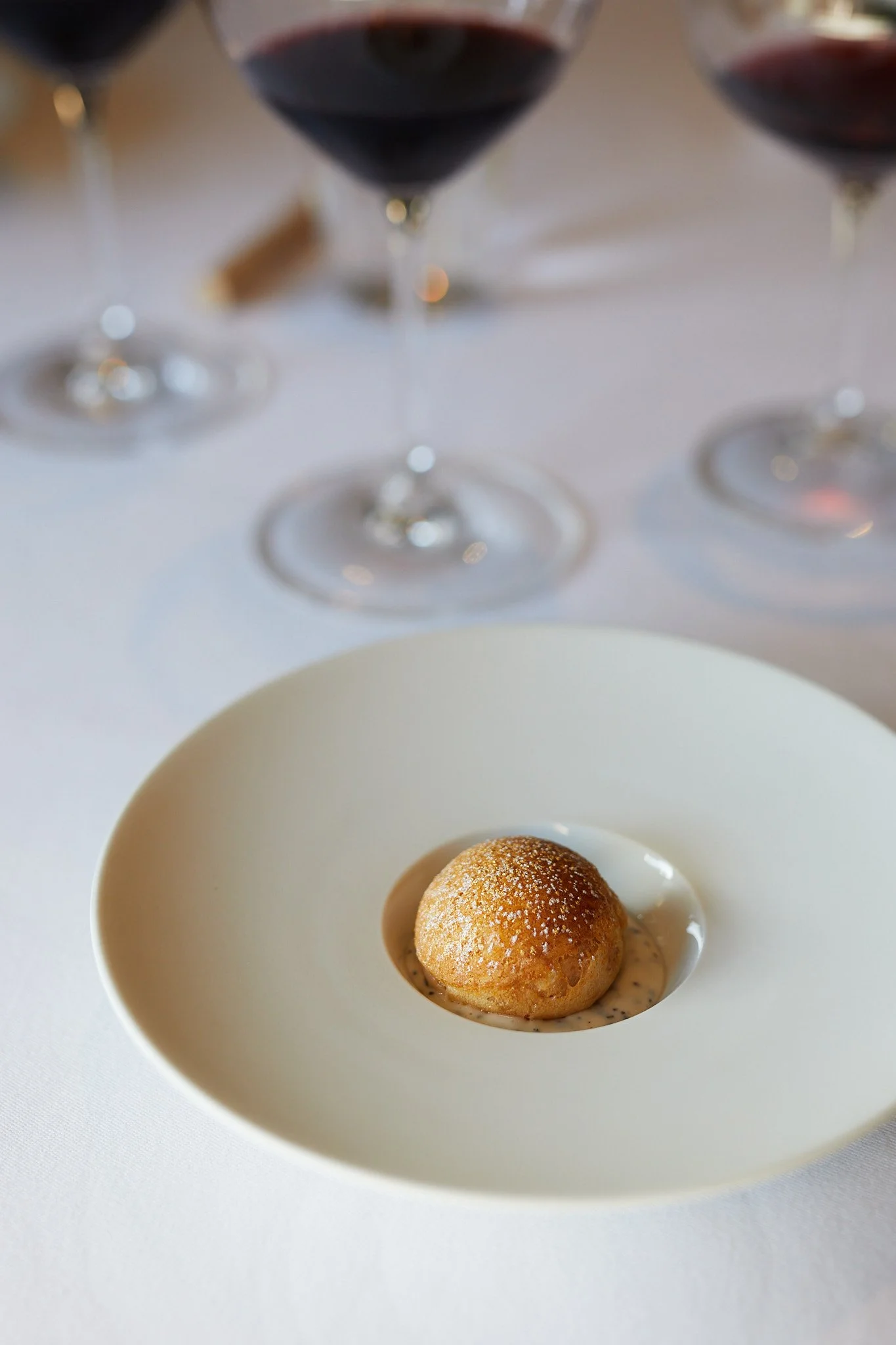 A small, round, golden brown appetizer on a white plate, garnished with a dusting of powdered sugar, with a creamy sauce underneath. In the background, there are three glasses of red wine on a white tablecloth  at the French Laundry.