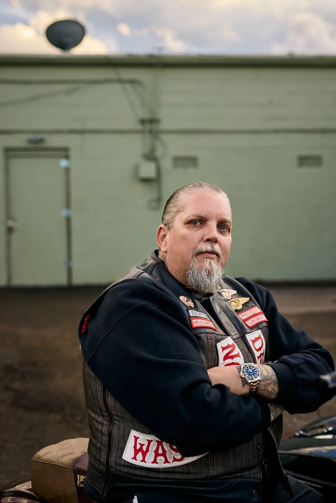 A middle-aged man with a beard and mustache, wearing a black jacket with patches and badges, is sitting on a motorcycle with his arms crossed, in front of a green wall.