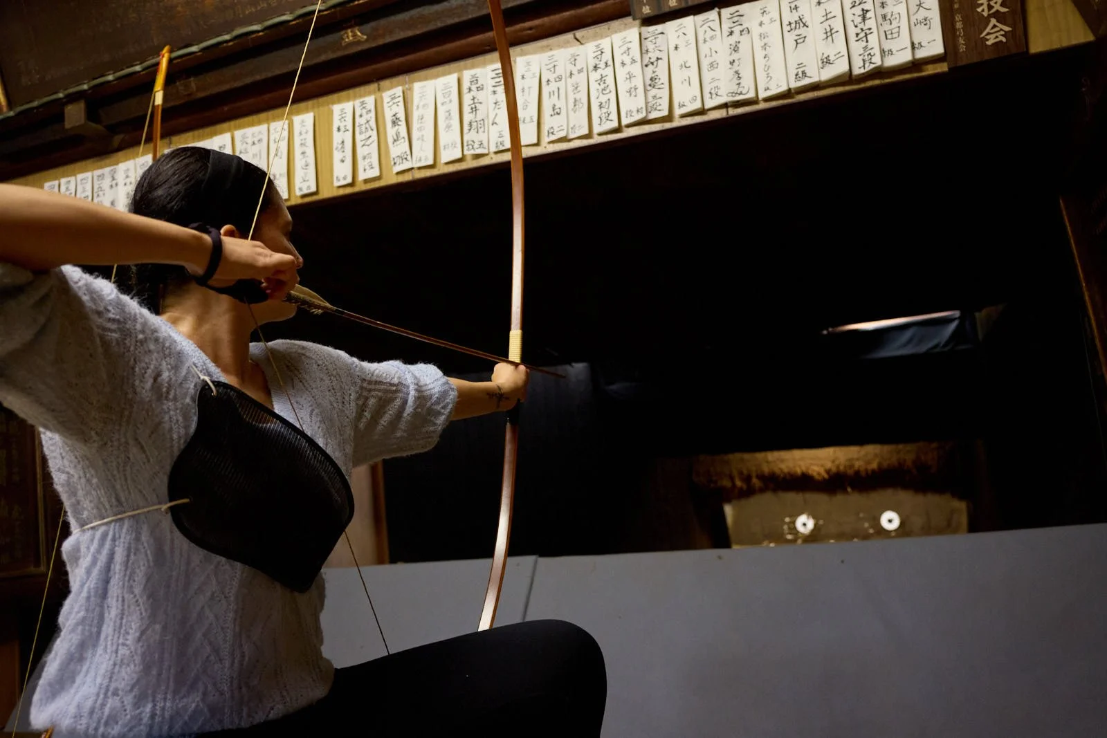 A woman practicing archery indoors, aiming with a traditional bow, in front of a wooden wall with Japanese writing plaques.