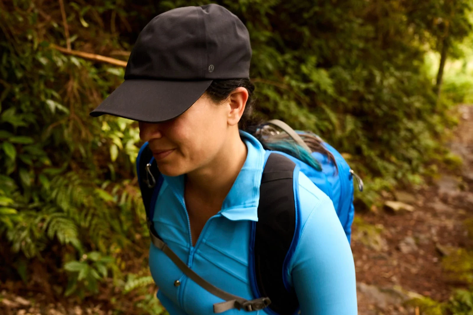 A woman hiking on a trail in Japan surrounded by green foliage, wearing a black cap, blue jacket, and carrying a blue backpack.