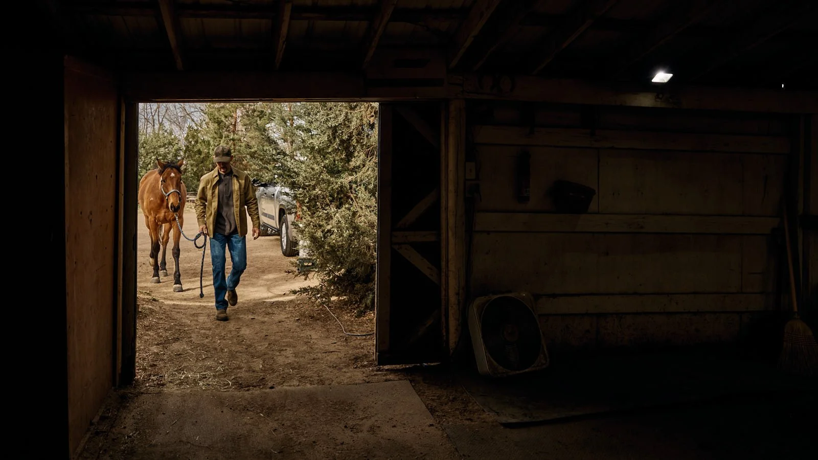 Man leading a brown horse out of a barn on a dirt path, with trees and a parked vehicle in the background.