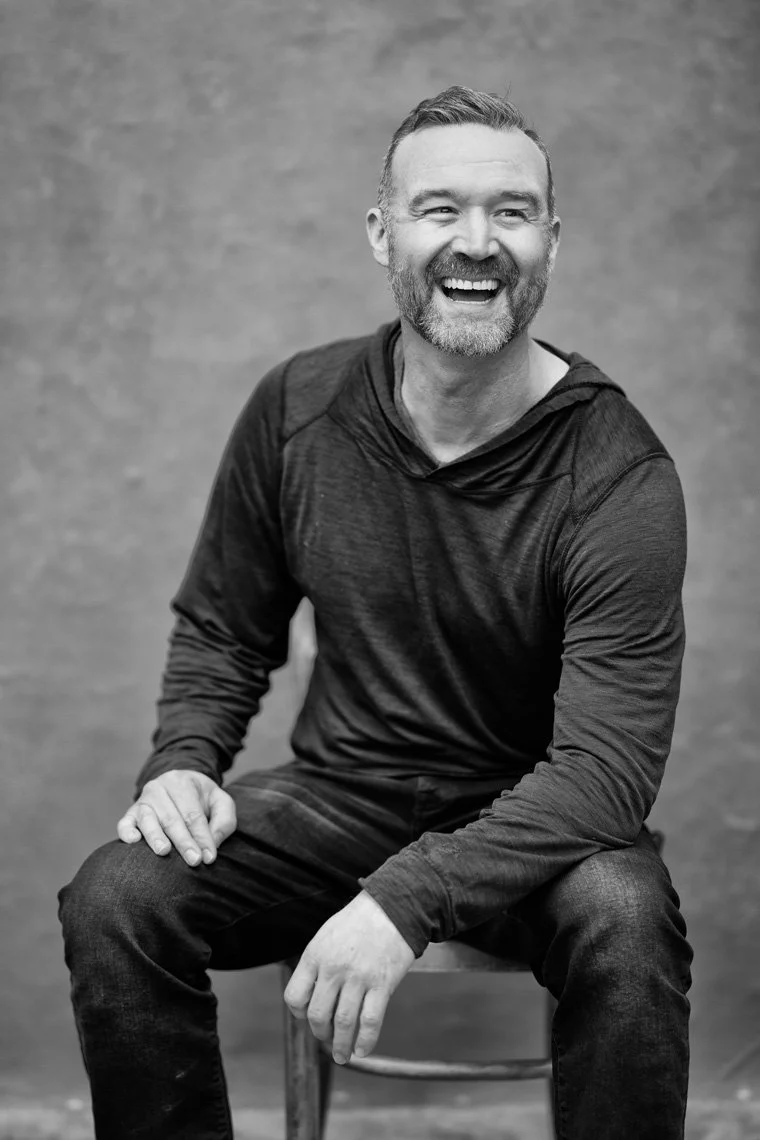 Black and white portrait of photography John Robson sitting on a wooden chair
