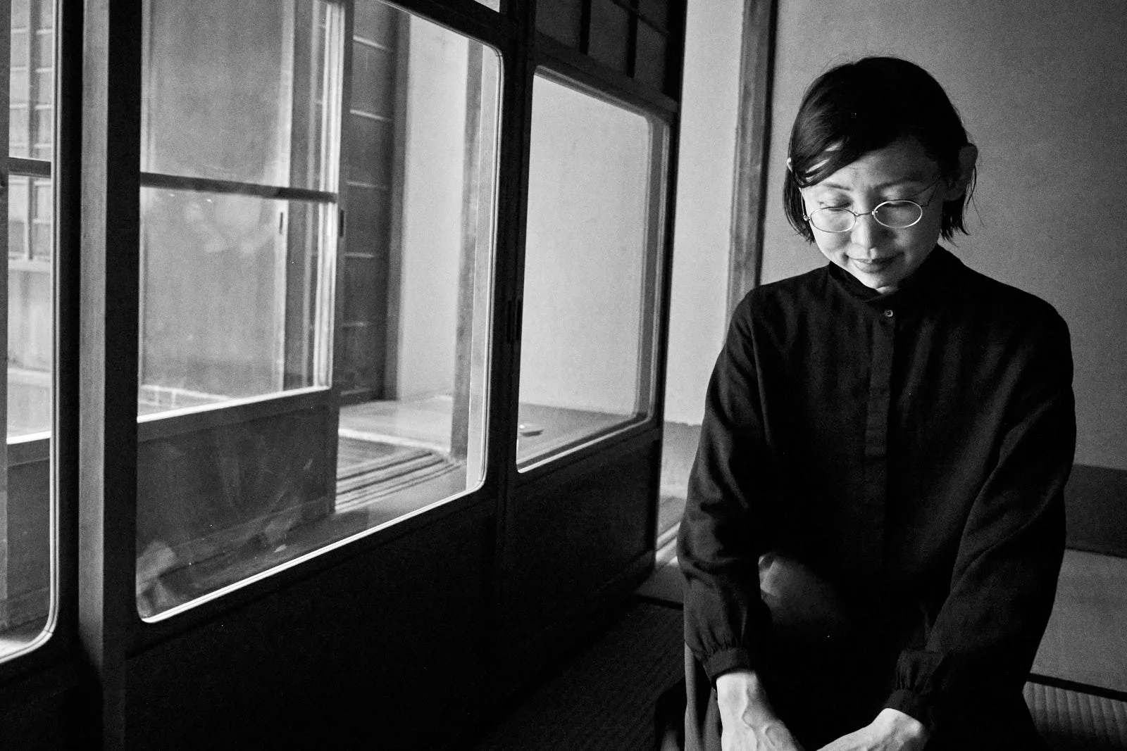 A woman with short hair, glasses, and a dark shirt sitting indoors near a glass display case, looking down with a gentle smile.