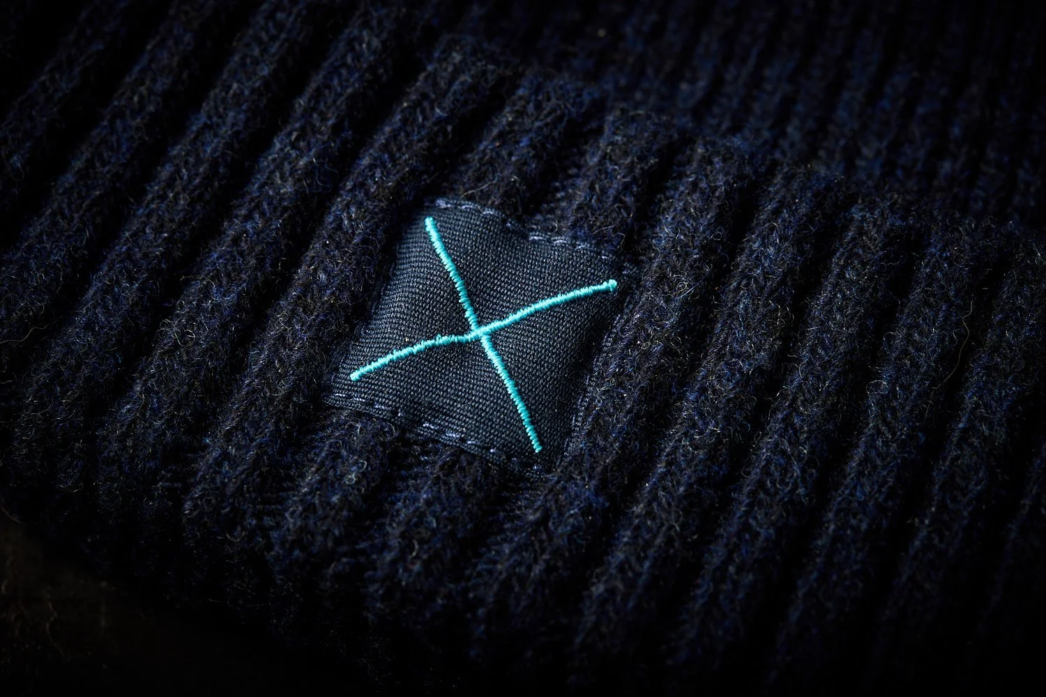 Close-up of a clothing label sewn into dark knitted fabric, featuring a light blue embroidered logo with two intersecting lines forming an X