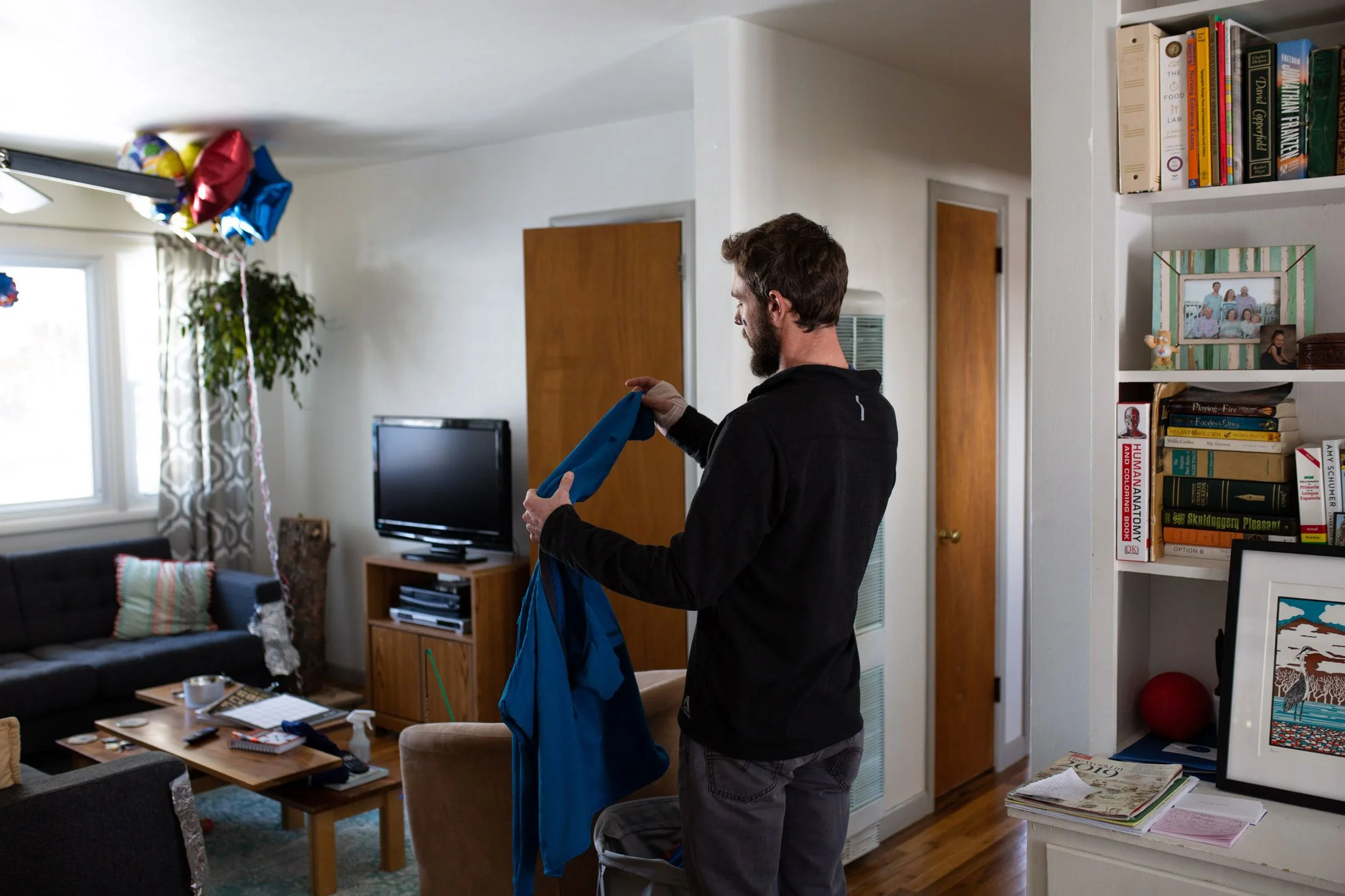 A man stands in a living room, holding a blue jacket, with balloons hanging from the ceiling near the window.
