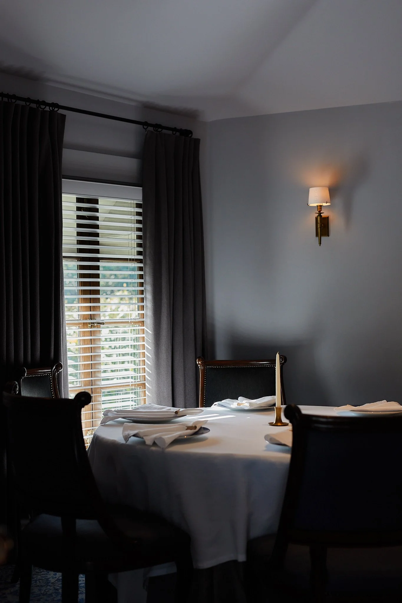 French Laundry dining room with a round table covered with a white tablecloth, set with napkins and a candle, surrounded by chairs, with a window with blinds and dark curtains, and a wall-mounted lamp on a light-colored wall.