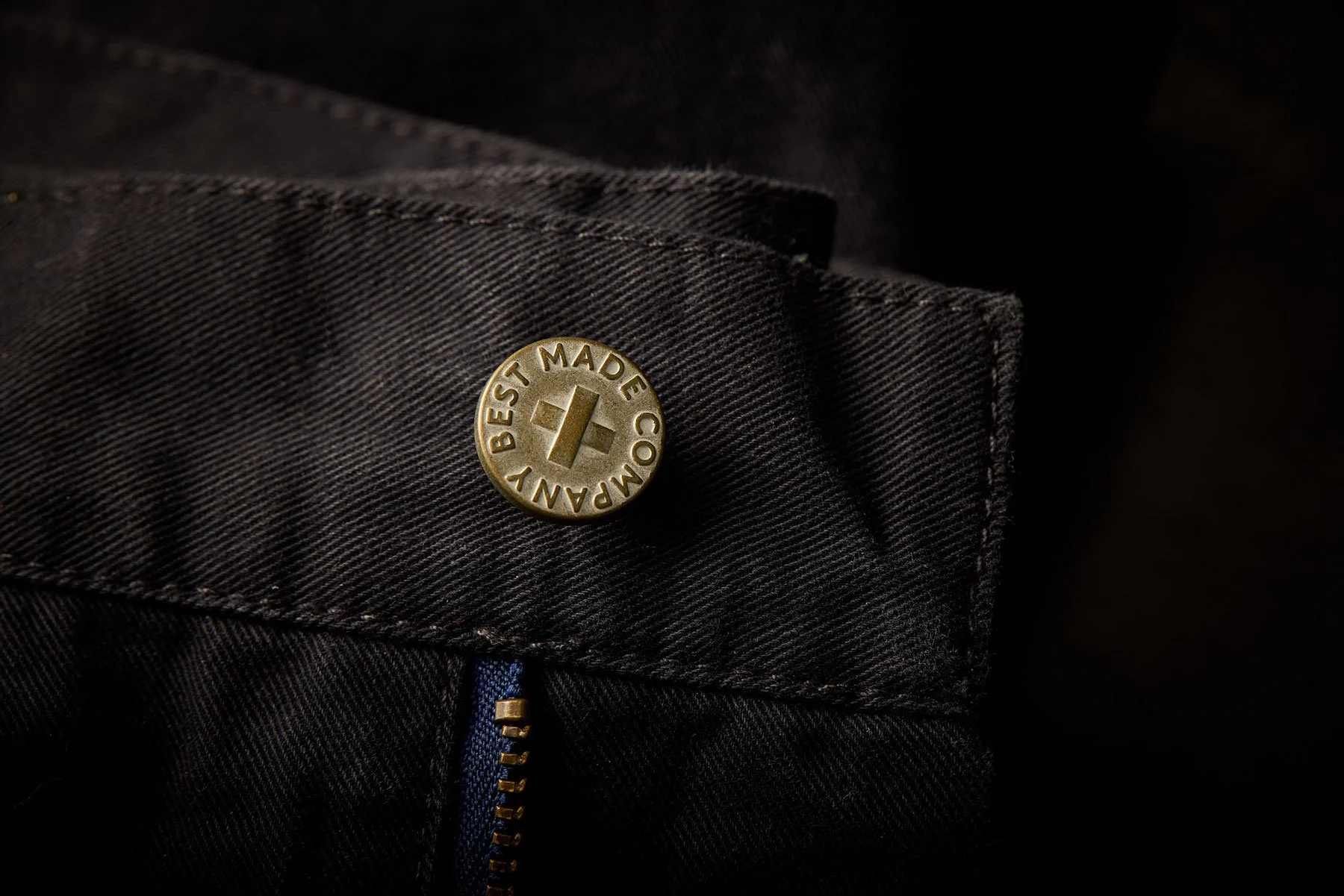 Close-up of a black fabric with a metal button that says 'Best Made Company' and a partially visible blue zipper.