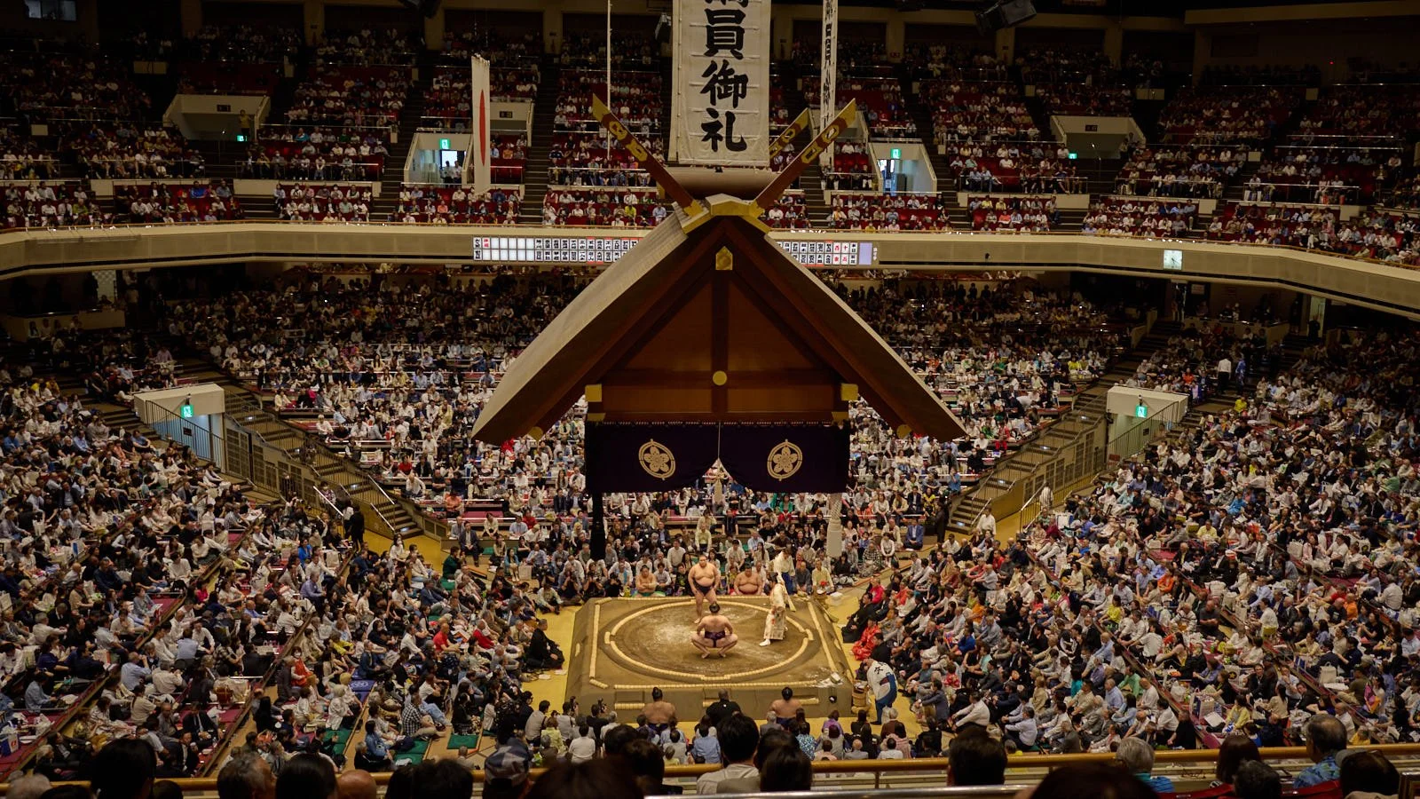 A sumo wrestling match taking place in a large indoor stadium with a big audience seated around the ring, which is at the center of the image. The ring is circular and elevated, with sumo wrestlers and a referee.