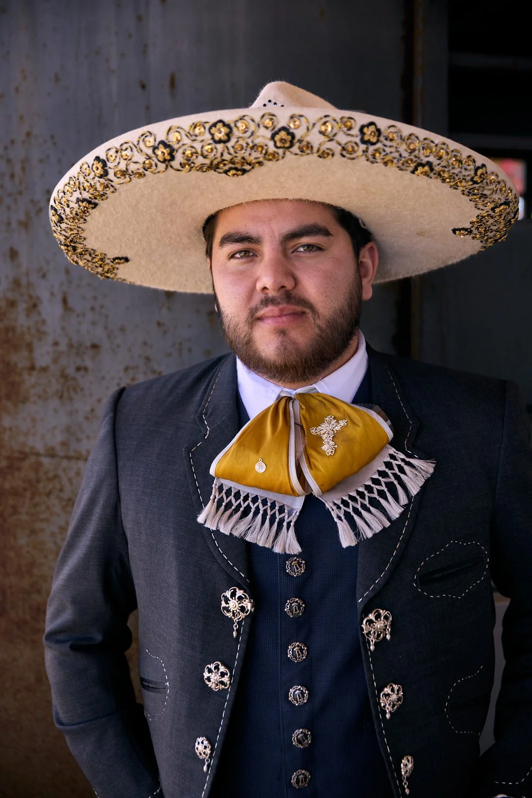 Man wearing a large sombrero hat, traditional mariachi attire, standing outdoors against a rusted metal background.