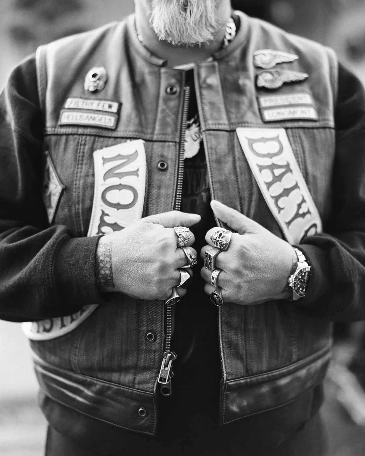 Close-up of a person wearing a denim jacket with patches and badges, holding the open zipper with both hands. They have rings on fingers and a watch on the left wrist.