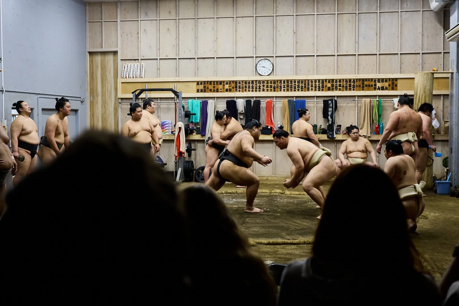 Sumo wrestlers practicing in a gym with wooden walls and a clock, spectators watching from the foreground.