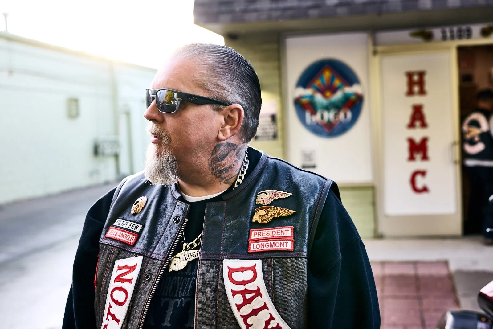 A man with sunglasses, a beard, and a tattoo on his neck, wearing a leather vest with patches, standing outside near a building with a sign that says 'HAMC'.