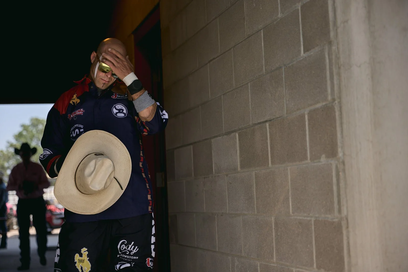 Rodeo clown in a colorful sports jersey standing against a brick wall, holding a straw hat in one hand and touching his forehead with the other,