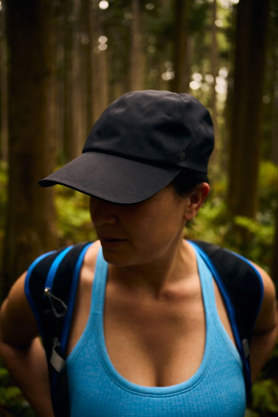 A woman in a blue Lululemon tank top and black cap with a backpack standing in a forest.