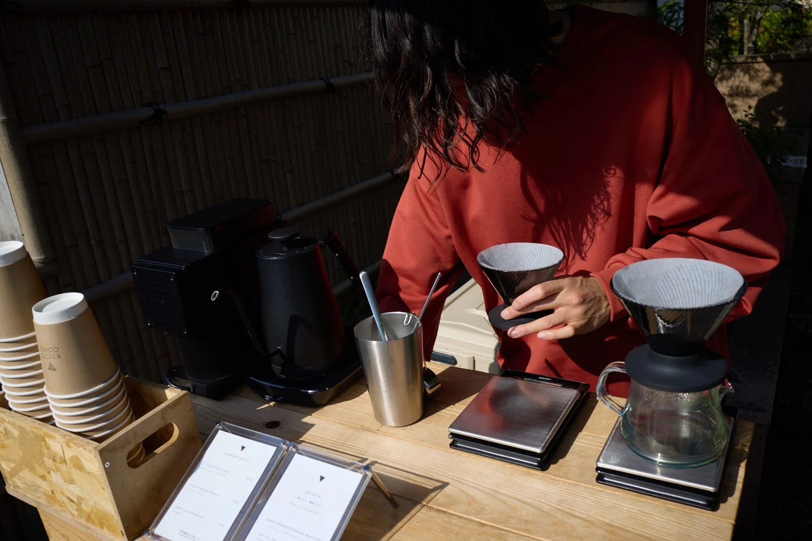 A barista preparing pour-over coffee at an outdoor coffee stand with black coffee equipment, cups, and a menu.
