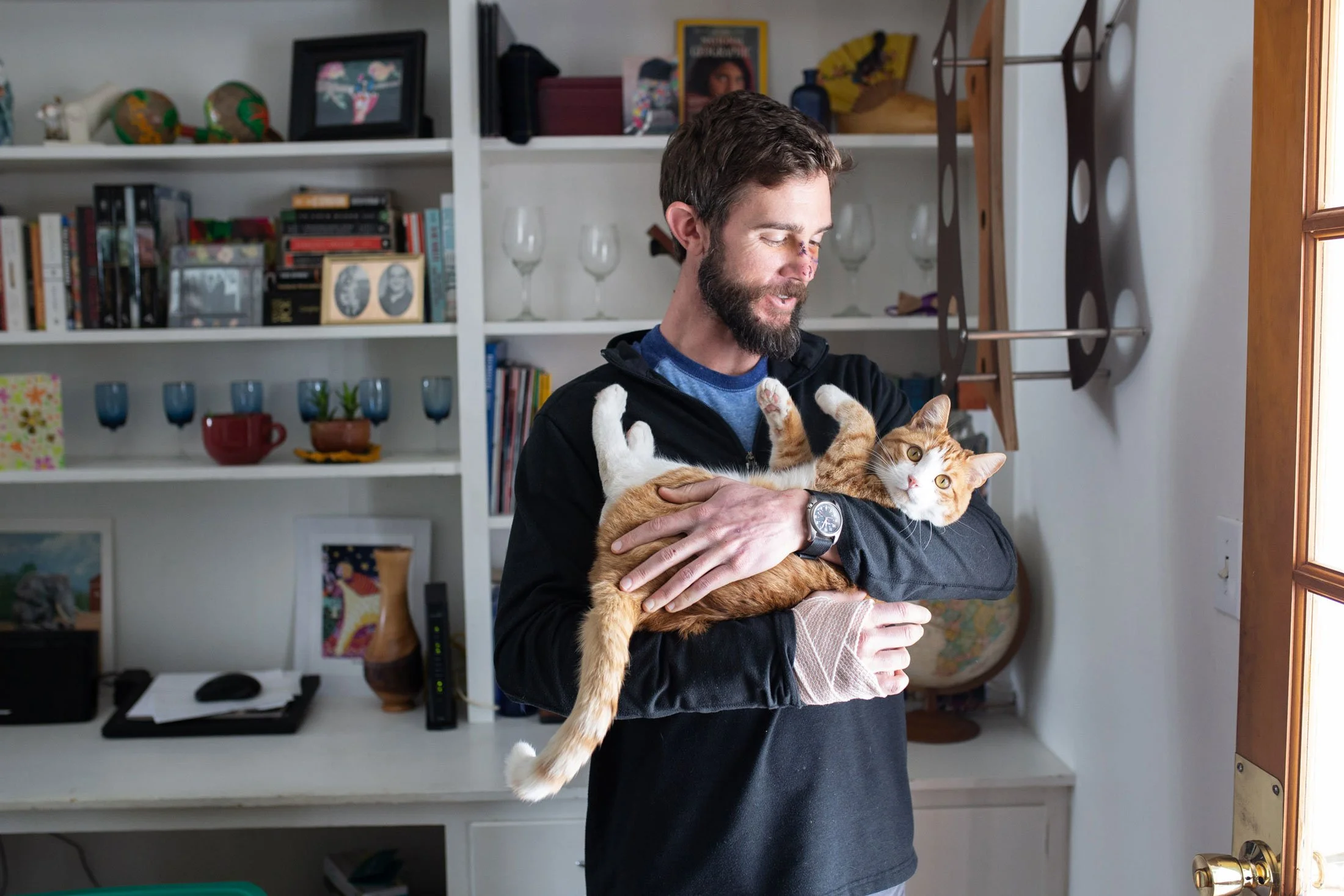 A man with a beard, wearing a black jacket, holds a ginger cat inside a room with shelves full of books, glassware, and decorative items. The man is smiling, and the cat is looking directly at the camera.