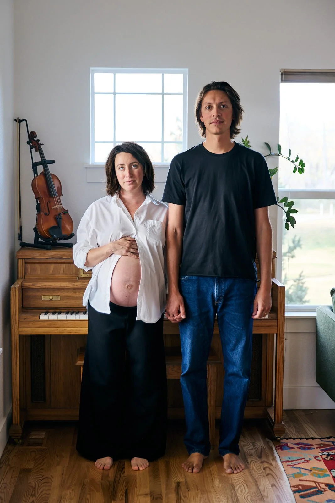 A pregnant woman and a man holding hands, standing in a room with a window, a violin on a stand, and a piano in the background.
