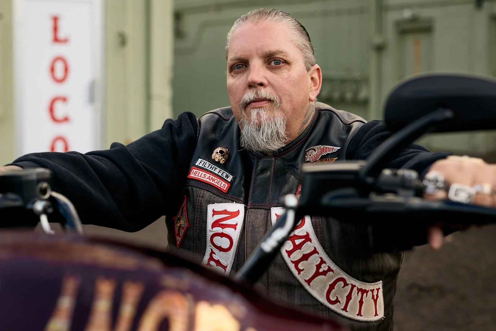 A man with a gray beard and combed-back hair wearing a black leather vest with patches sits on a motorcycle, looking directly at the camera. The background shows a green building and a white pole with red text.