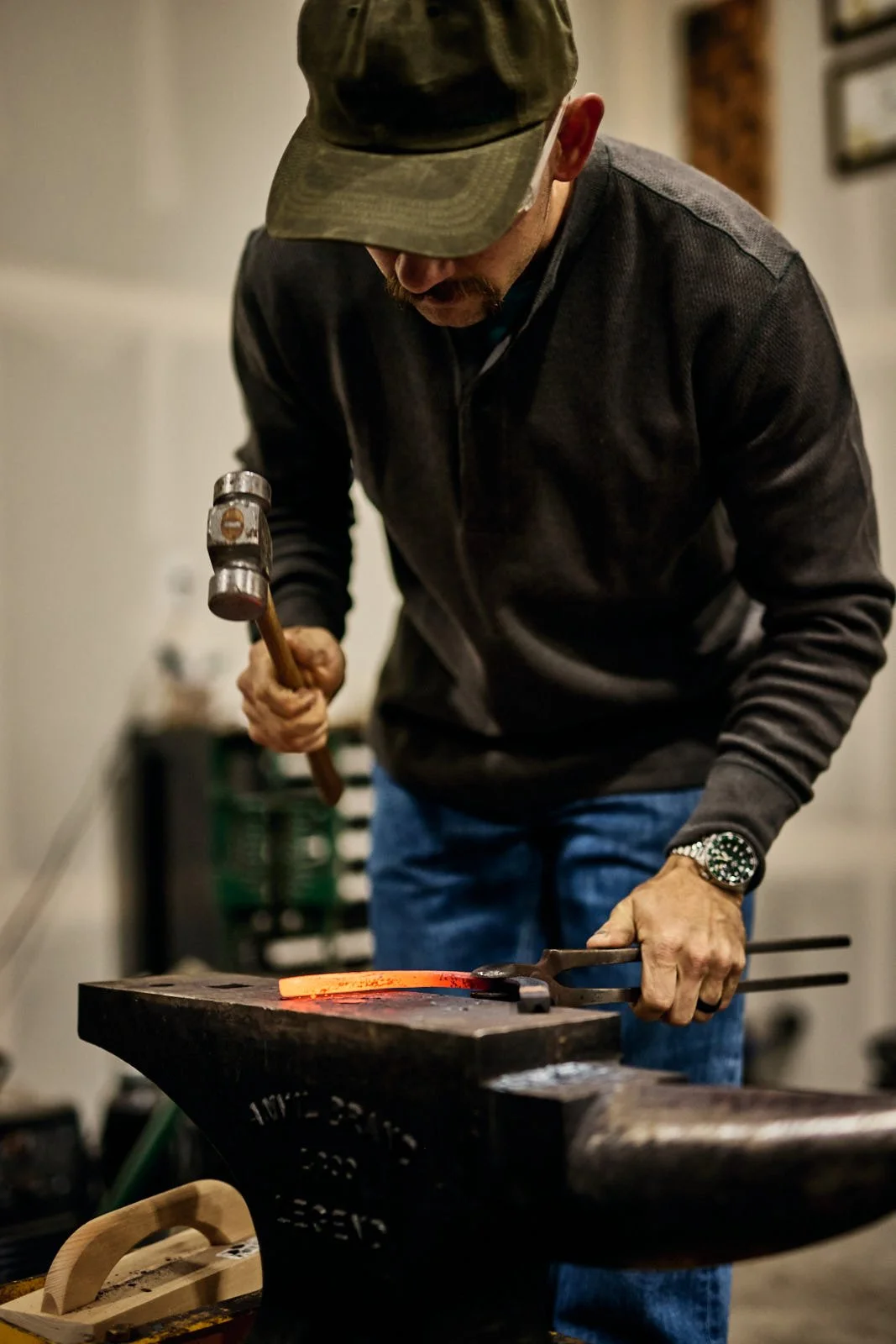 A man wearing a cap and a dark jacket hammers a glowing horseshoe on an anvil in a workshop.