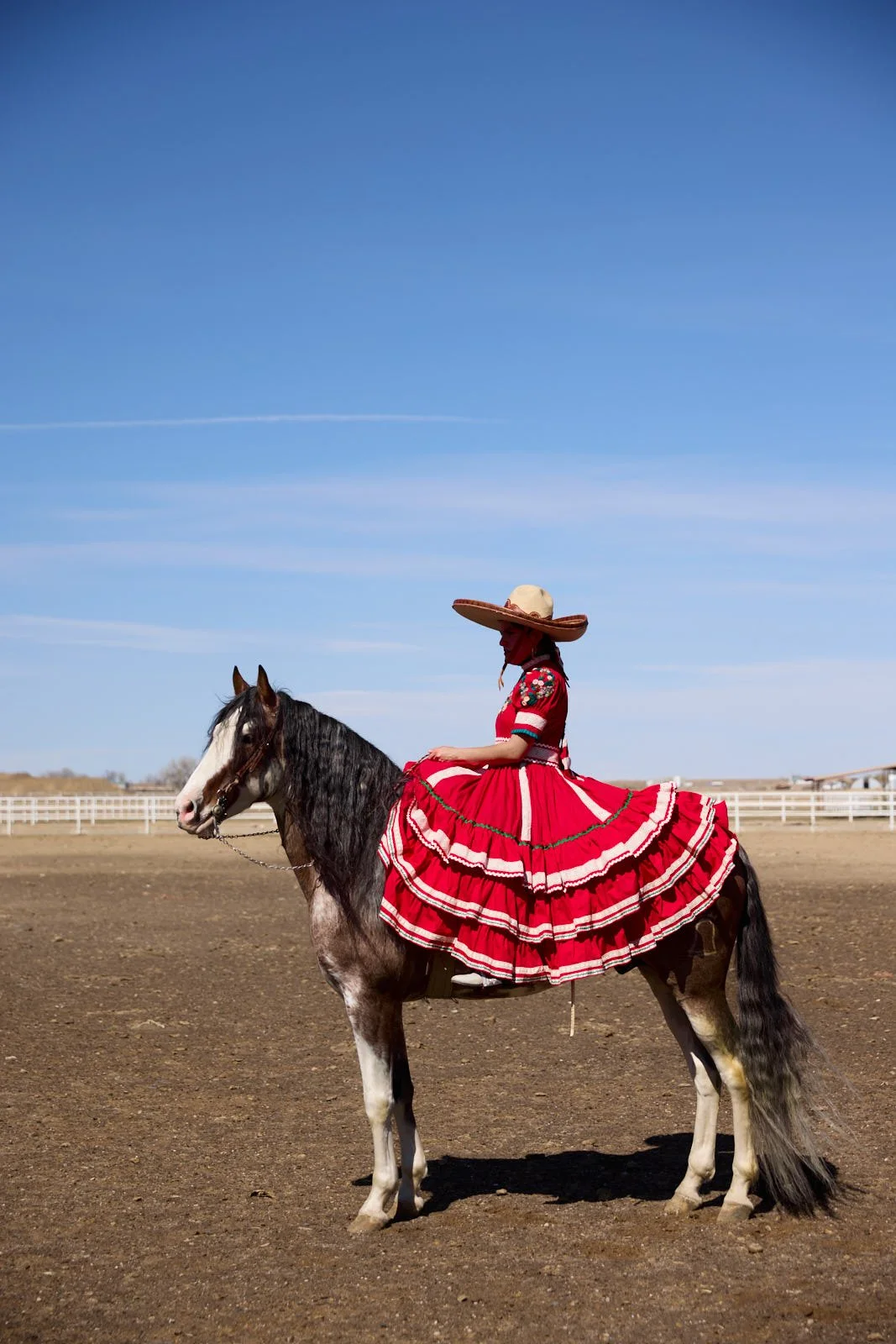 A woman riding a horse in an open field, wearing a wide-brimmed hat and a vibrant red dress with ruffles, under a clear blue sky.