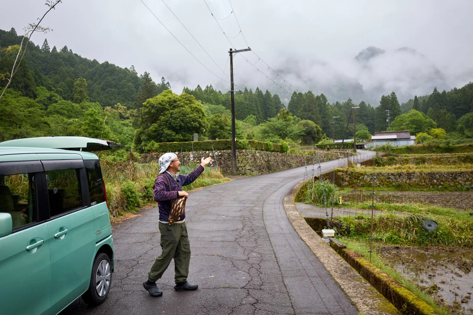 A person standing next to a parked car on a rural mountain road, reaching out towards the foggy, tree-covered mountains in the background, with cultivated fields and utility poles alongside the road.