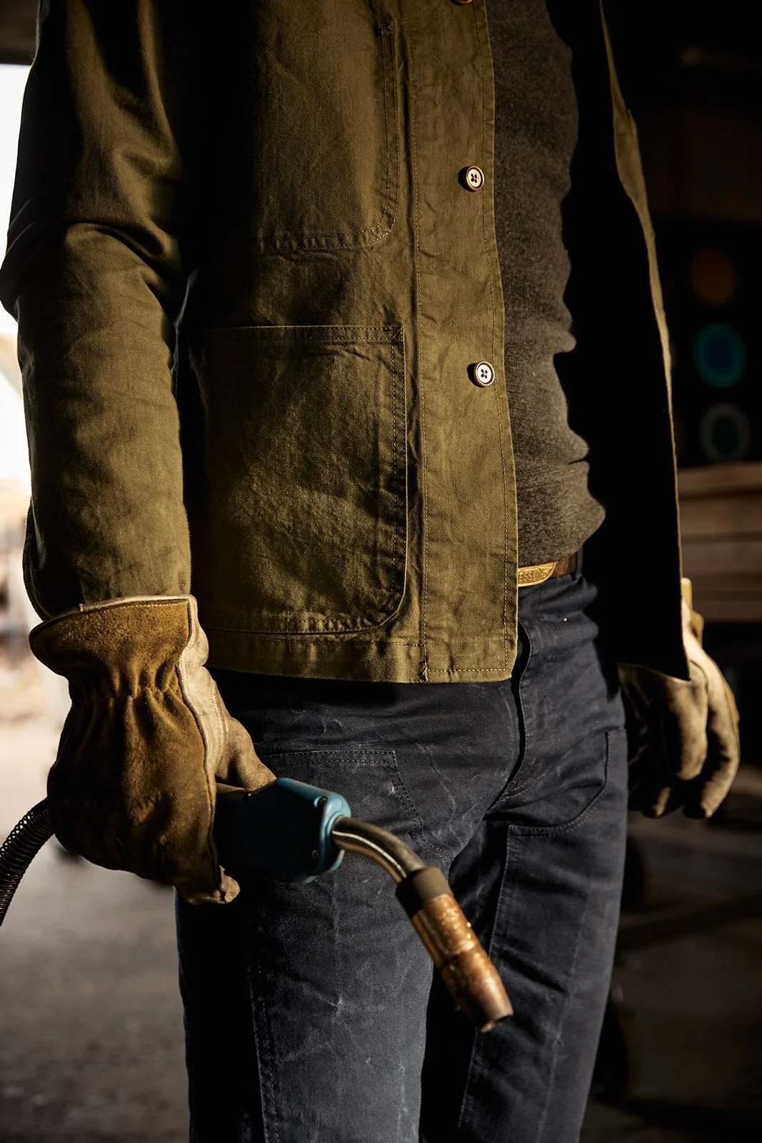 A person wearing safety gloves holding a welding torch, dressed in a green jacket, black shirt, and black jeans, in an industrial setting.