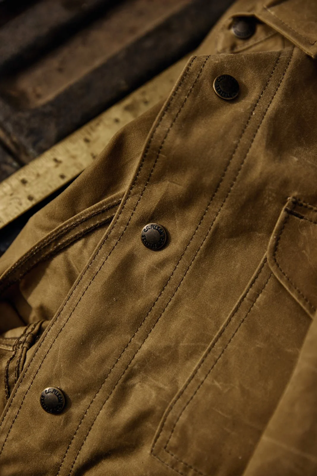 Close-up of a brown jacket with black metal buttons, showing detailed stitching and a pocket, laying on a rustic wooden surface.