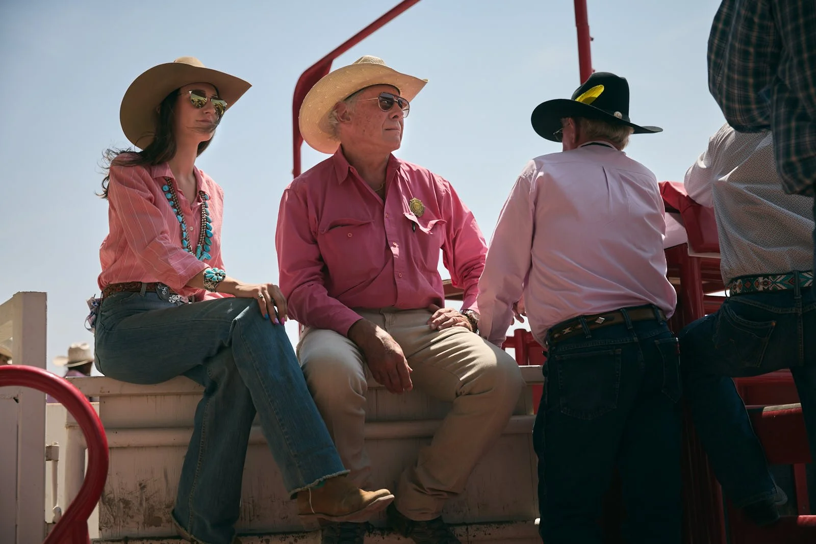 Group of people dressed in Western attire sitting and standing on a cattle trailer outdoors under a clear sky.