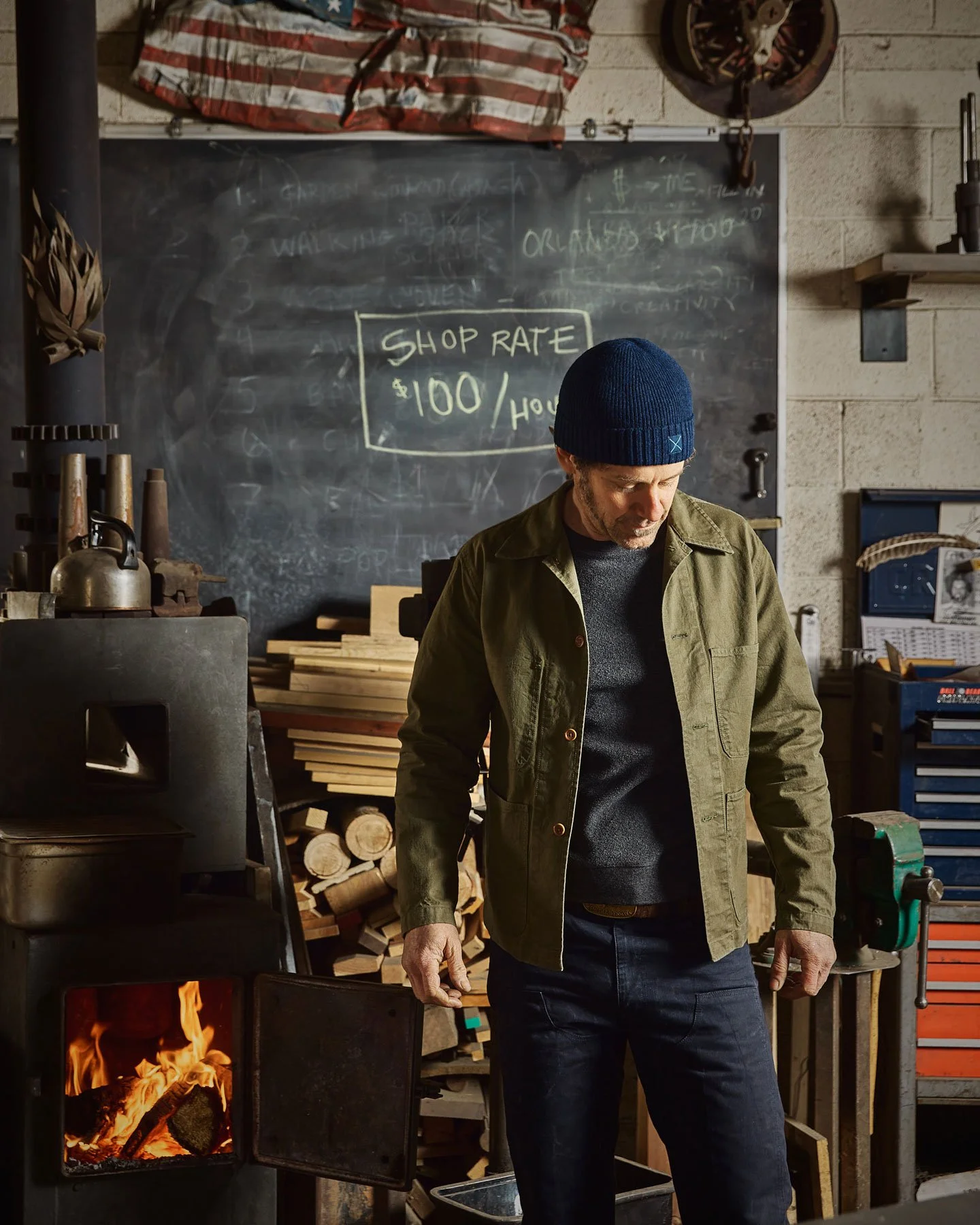 Man wearing a blue beanie and green jacket looking down in a woodworking workshop with a blackboard behind him that reads 'Shop Rate $100/Hour'.