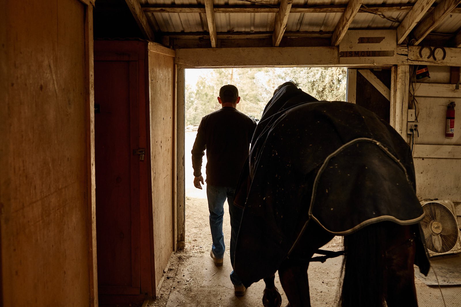 A man walking out of a barn or shed with a horse nearby, inside a rustic wooden structure with a fan and a fire extinguisher visible.