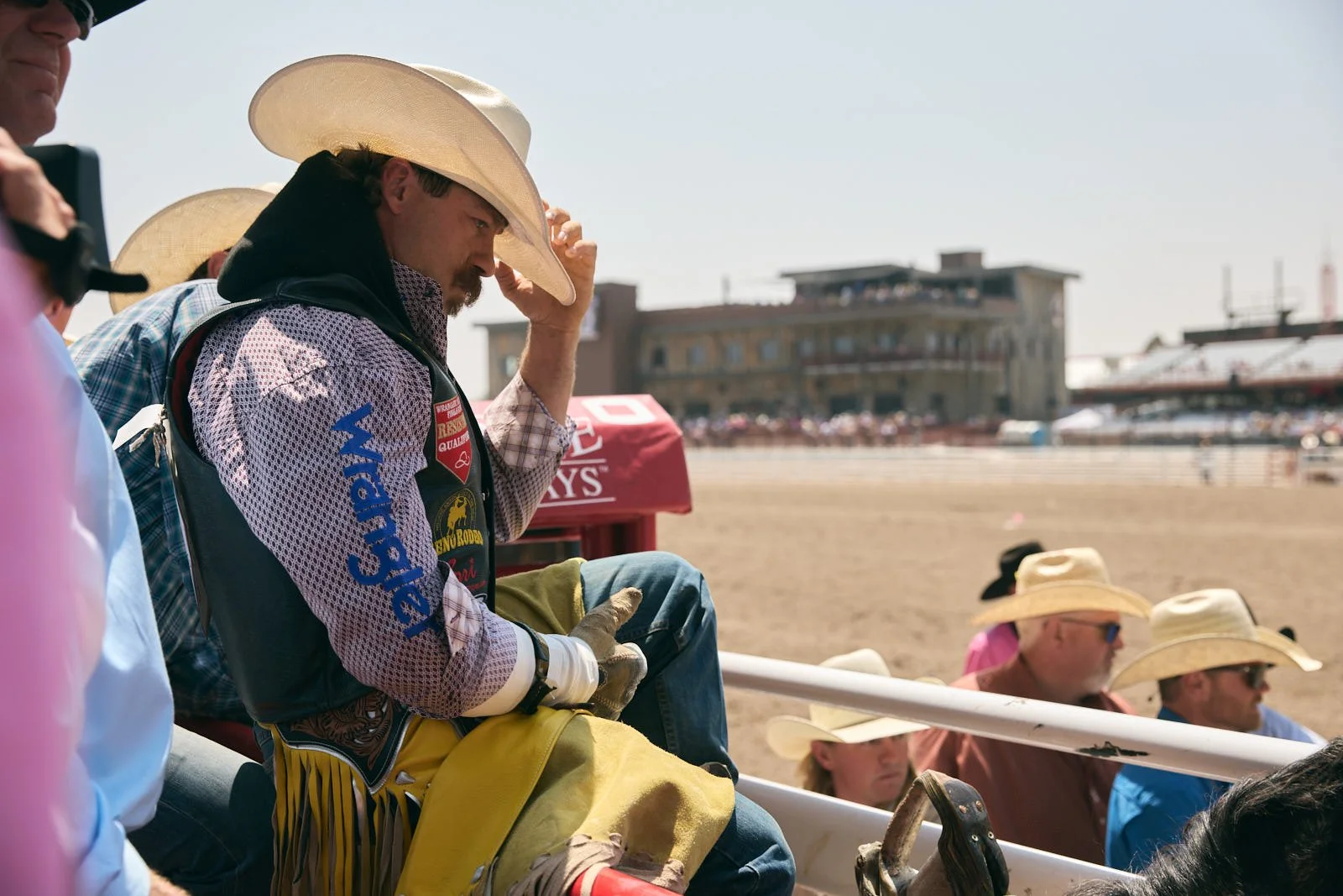 Man in cowboy hat preparing to ride a bull with other spectators, some also wearing cowboy hats, and grandstand in background.