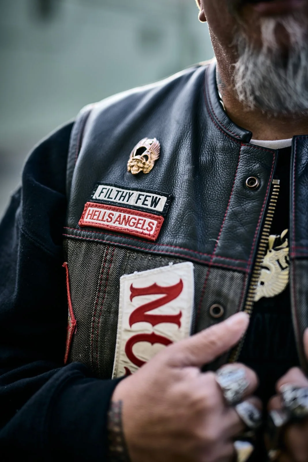 Close-up of a person wearing a black leather vest with patches that say along with a skull pin and a badge. The person has rings on their fingers, a beard, and tattoos visible on their hand.