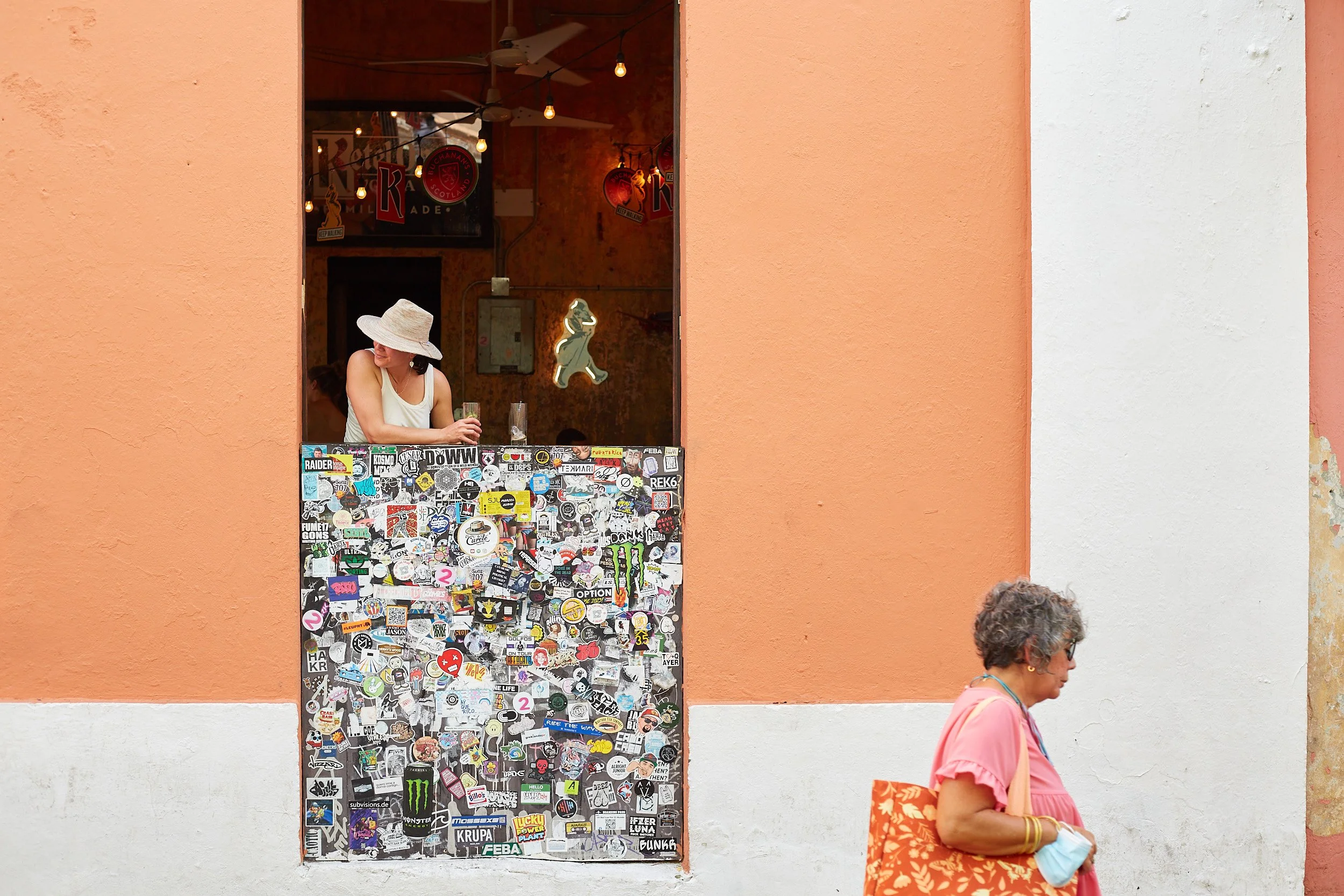 A woman wearing a large hat and white tank top looking out from a window decorated with various stickers, with another woman walking past in the foreground on a pastel-colored wall.