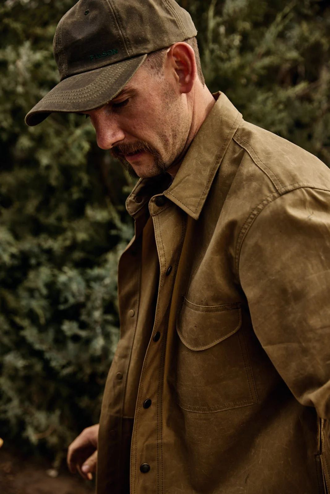 A man wearing a brown cap and a brown jacket, looking down in an outdoor setting with blurred greenery in the background.