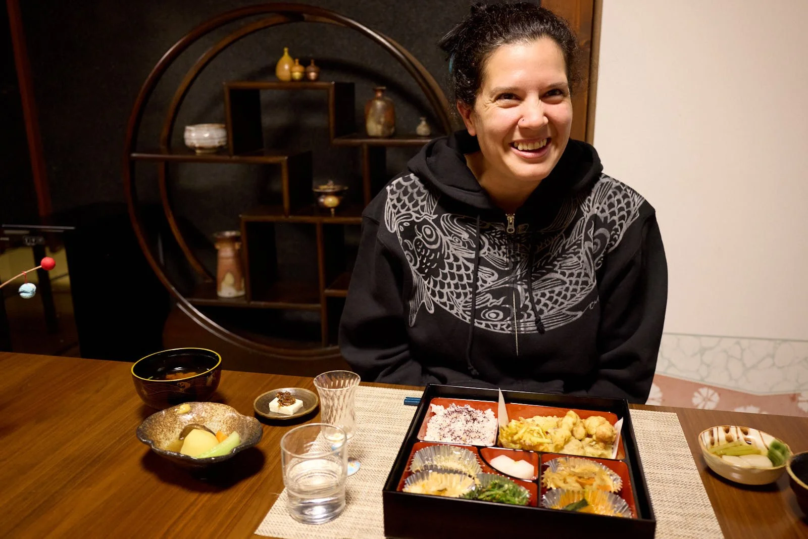 Woman with black hair and a black hoodie with a white pattern, smiling, sitting at a table with a Japanese meal set in a black and red box, various small dishes, bowls, and glasses of water around her.