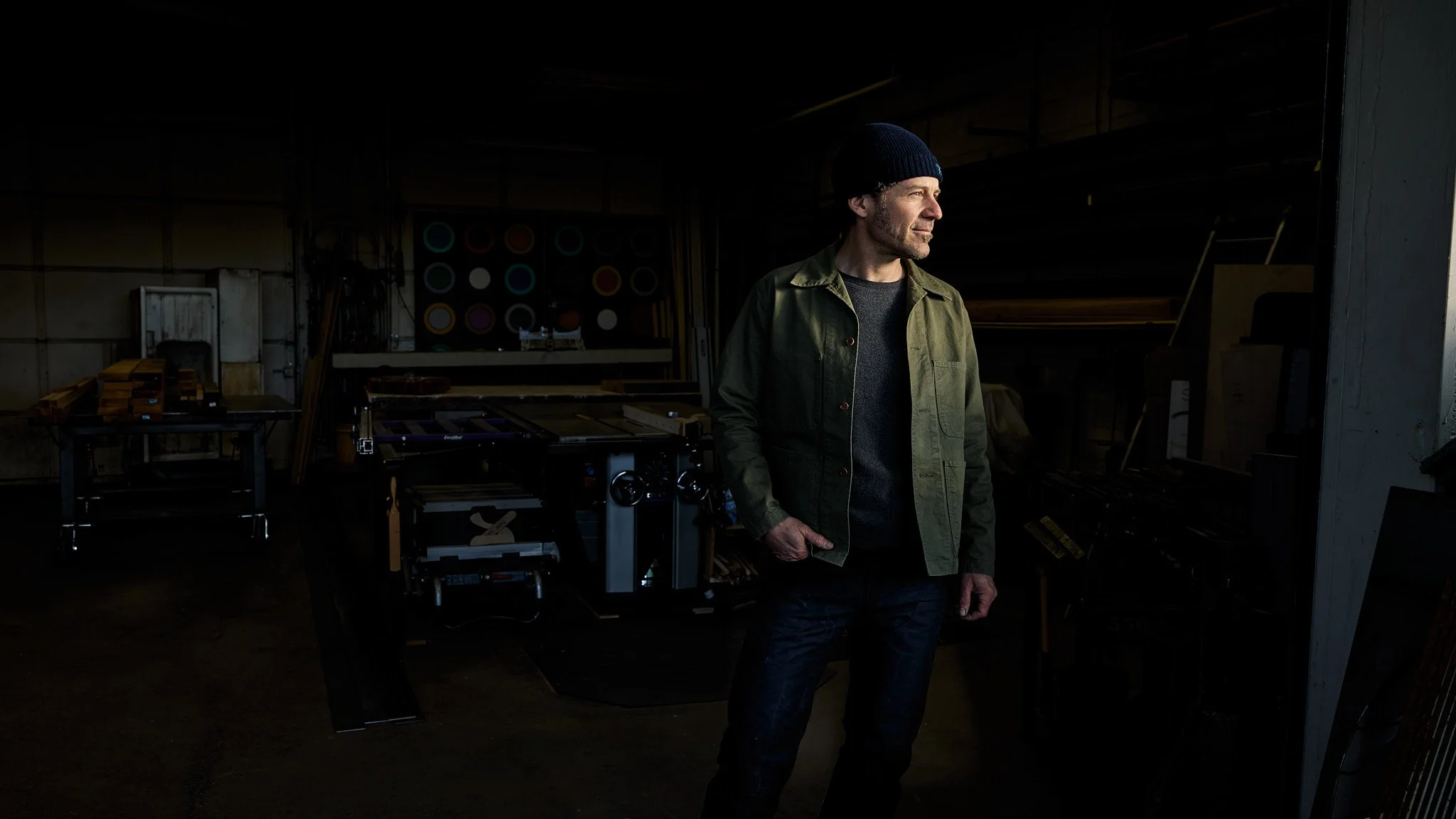 A man wearing a navy blue beanie and olive green jacket standing inside a dimly lit workshop