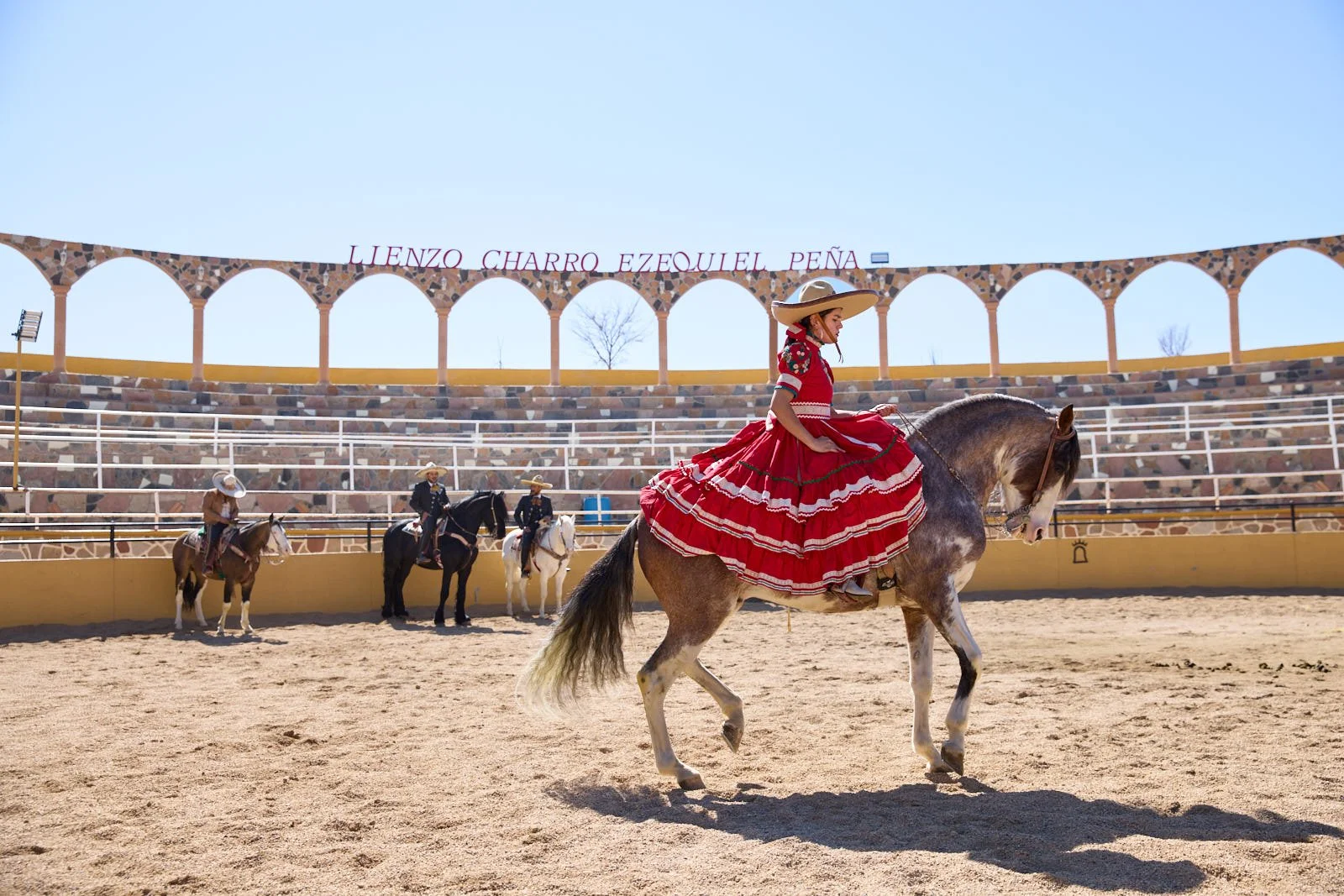 A woman in a traditional Mexican dress riding a horse in a rodeo arena, with three other riders on horseback in the background, and a sign with the names 'Llenzo Charro Ezequiel Peña' in the stands under a clear blue sky.