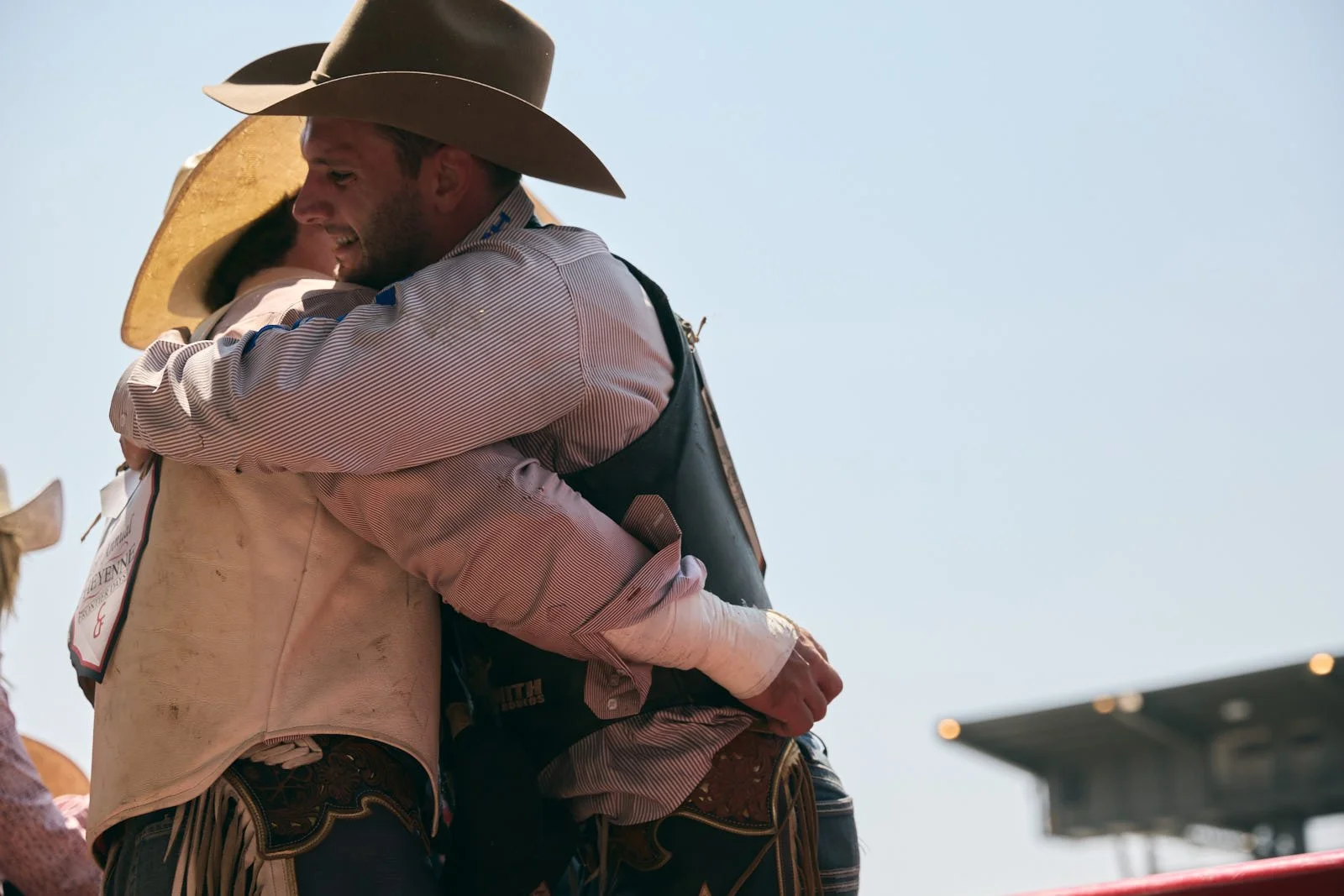 Two men in cowboy hats hugging each other at an outdoor event on a sunny day.