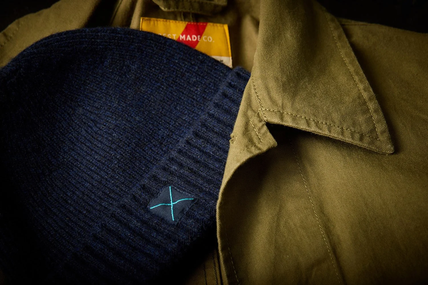 A close-up of a navy blue knit beanie hat with a small black and turquoise logo patch, placed on an olive green collared shirt.