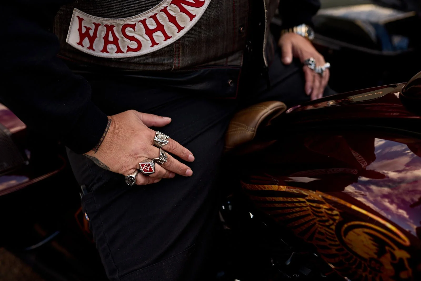 Close-up of person wearing multiple rings, sitting on a motorcycle with a reflective, colorful surface. The person has tattooed arms and is wearing a black shirt and a vest
