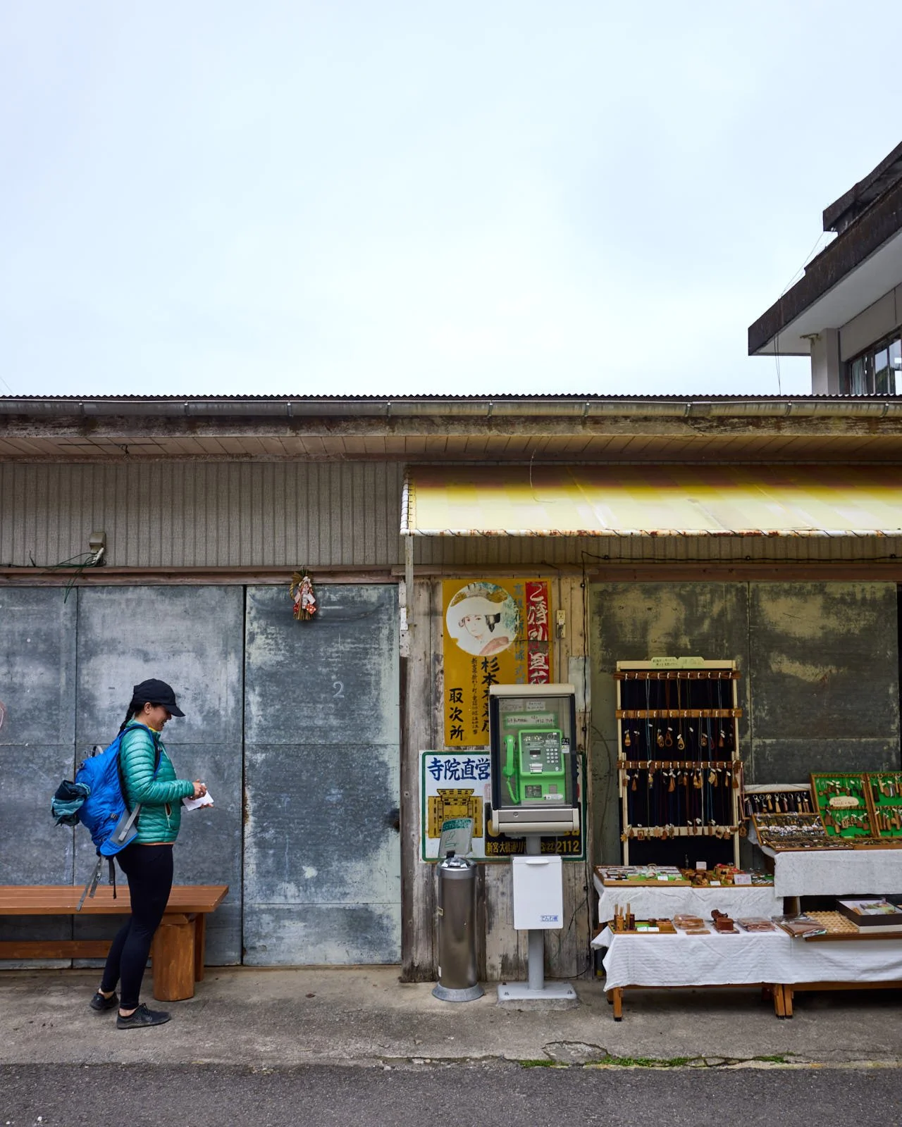 A woman with a backpack and a hat standing outside a small shop with jewelry and souvenirs, a payphone, and Japanese signs.