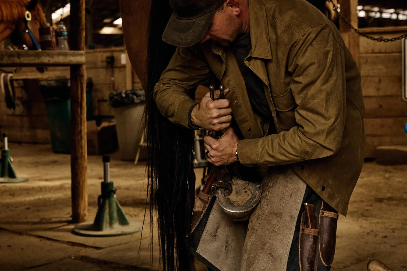 Man in a brown jacket and cap working in a horse barn, holding a piece of equipment.