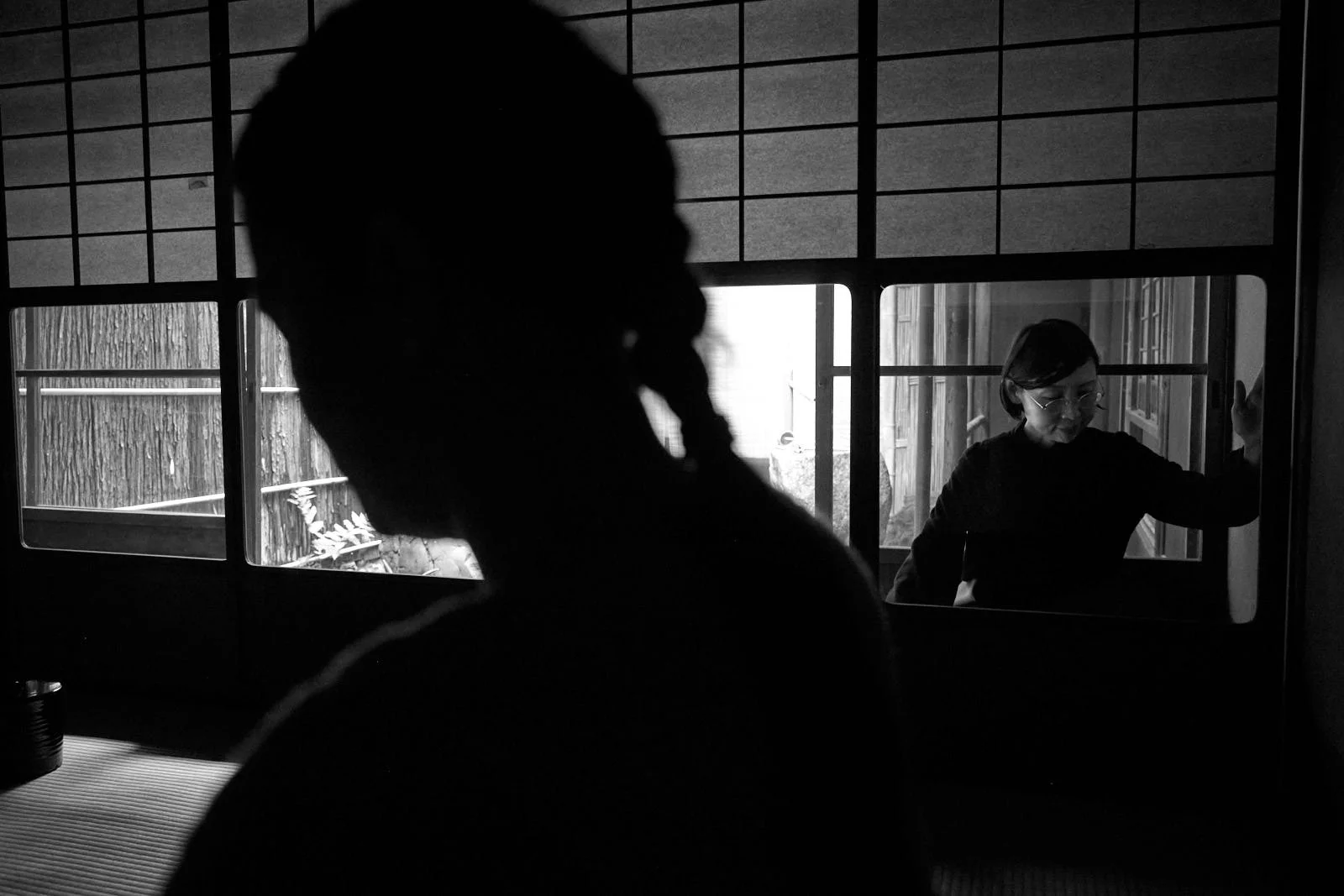 A black and white photo showing the silhouette of a person in the foreground, with a woman wearing glasses in the background, seen through a window or glass panel.
