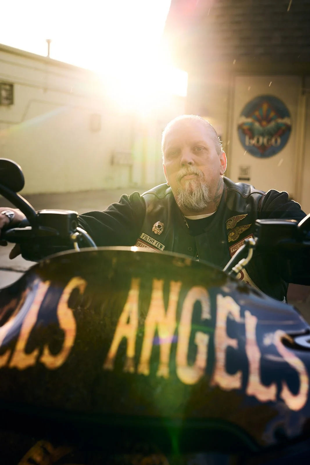 A man with a beard and long hair, dressed in a leather vest with patches, sits on a motorcycle with a Harley Davidson logo on the front. The background is sunlit with the sunlight shining behind him.