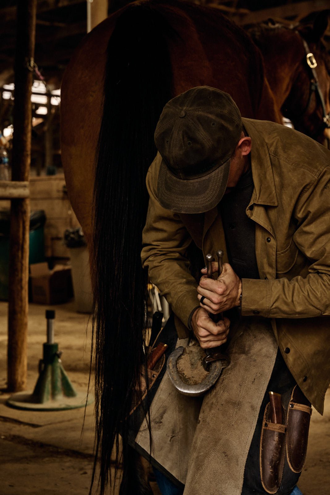 A person wearing a cap and apron working with tools on a horse's hoof inside a barn