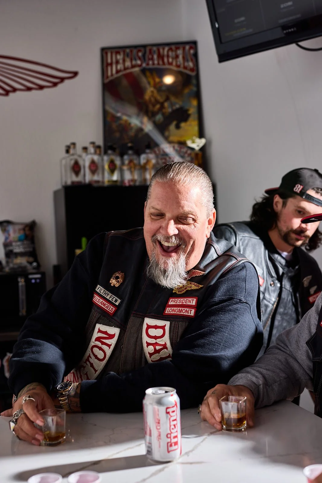 A group of men sitting at a table, with one man smiling and holding a glass of alcohol, in front of a backdrop with a poster and several bottles, with a TV screen mounted on the wall.